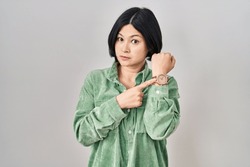 Young asian woman standing over white background in hurry pointing to watch time, impatience, looking at the camera with relaxed expression 