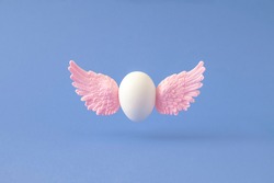 White Easter egg with pink angel wings flying against blue background. Creative resurrection concept.