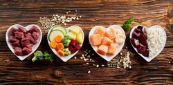Panorama banner of healthy fresh ingredients for pet food in individual heart-shaped bowls viewed from overhead with chopped raw beef, liver and chicken , mixed vegetables and rains on rustic wood