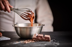Chef whisking melted chocolate in a stainless steel mixing bowl using an old vintage wire whisk in a close up on his hand