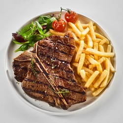 Tasty grilled or barbecued T-bone steak with fried potato chips, salad greens and roasted tomato viewed from above over white in square format