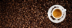 Fresh roasted coffee bean background with a cup of hot frothy espresso placed to the right side in a wide panorama banner with copy space