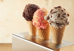 Three ice cream cones with assorted flavors including chocolate, strawberry and choc chip standing in a metal holder on a counter