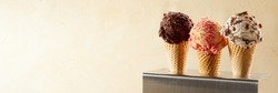 Three servings of tasty creamy ice cream in cornets with a scoop of strawberry, chocolate and Oreo or choc chip against a neutral beige gradient background with copy space in a panorama banner