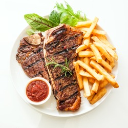 Grilled t-bone or porterhouse steak seasoned with rosemary and served with golden French fries, fresh leafy herb salad and a tomato dip, high angle view on white