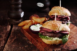 Gourmet Tasty Steak Burgers with Ham Slices on a Wooden Tray with Potato Wedges and Dipping Sauce.