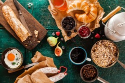 Wholesome rustic breakfast with cereal, fried egg, bread, jams, sausage, milk in a can and coffee spread out on a green wooden background, overhead view