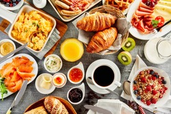 Large selection of breakfast food on a table with muesli , eggs, croissant, orange juice, fruit, smoked salmon, cold meats, cheese and jam served with coffee viewed from above