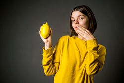 Funny girl in a casual yellow sweater holds a lemon fruit in the hand. She is thinking about the benefits of lemons and vitamin C. The woman is worried about an unsuccessful purchase - to buy a lemon.