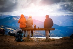 Group of young travelers sit on a wooden bench and enjoy a stunning sunset in snow-capped mountains from a view point. Man and two girls with backpacks and hiking equipment walk in the national park.