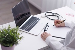 A woman doctor sits at a table looking into a laptop and making notes on paper. Medical background with doctor, laptop, stethoscope. Working in a hospital with a patient. High quality photo