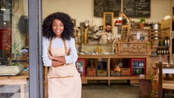 Portrait of a smiling young African American barista leaning with her arms crossed on the door of a trendy cafe 