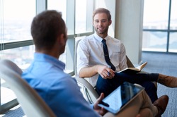 Young businessman sitting comfortably in an open modern office, smiling while having a positive conversation with a coworker