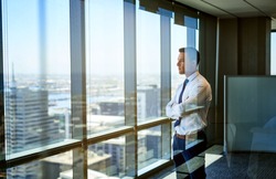 Confident young businessman deep in thought while looking through windows at the city from high up in an office building
