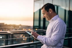 Smiling mature businessman working online with a digital tablet while standing outside on an office building balcony overlooking the city at dusk