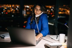 Young businesswoman talking on the phone and using a laptop at her office desk late into the night in front of windows overlooking the city 
