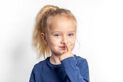 A little girl with blue eyes cheerfully touches her nose with her finger.