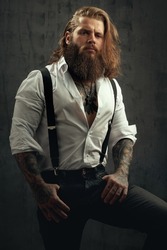 Handsome courageous middle-aged man with long hair and a thick beard posing in the studio in jeans and a shirt with suspenders