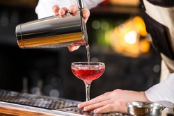 Male bartender is making cocktail pouring alchohol from shaker to glass at bar background.