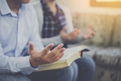 Close up of a man holding bible and raise hand up  praying to God with his friends while sitting on sofa at home, Christian family worship concept, copy space
