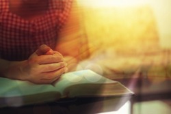 Close up of a woman hands praying on the open holy  bible on a table indoor with the windows light lay warm tone . Christian faith and trust concept  with copy space. Christian devotional background.