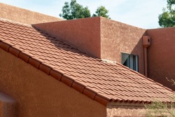 Long slanted adobe style roof with red tiles and orange stucco exterior with back and front yard trees in a neighborhood. In suburban area in late afternoon sun with desert design and facade.