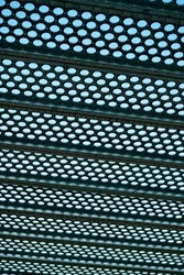 Black metal grate or drain vent with holes in geometric pattern for background or texture image with background sky. In industrial area with light pattern and horizonal beams in city.
