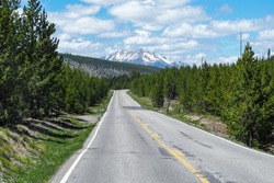 Road leading through the forest of Yellowstone National Park, Wyoming, USA, with trees all around and dramatic snowy peak in the far back. Infinity point road in the wilderness.