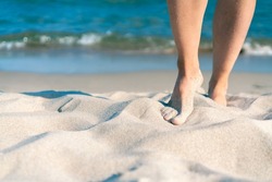 Bare female feet on a sandy beach with blue sea in the back on a beautiful sunny summer day. Concept for holiday, vacation, relaxation, mental health, joy, recreation.