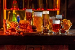 Glass of beer on bar counter. Jugs, mugs, pints of brew beverage, ale, cider, snack on wooden table in pub, bar. Backlit dark showcase with craft beer bottles in vintage brewery. 