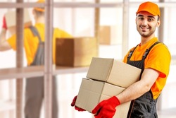 Young man holding cardboard package working in warehouse among racks and shelves. Delivery man with box. Staff laborer, orange uniform cap, t-shirt, coveralls service moving delivering orders goods. 