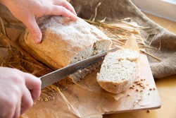 Woman slices fresh bread. hands with a knife. Freshly baked rye bread on a wooden board. Burlap, craft paper and straw on the background. Organic bread baked at home from wheat flour. Sliced bread.