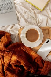 Home office workspace with office supplies: laptop, sweater, cup of coffee with milk, glasses, book and wood board on a messy bed. Fall, autumn concept.