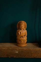 Hand carved wooden buddha figurine on natural wood shelf in front of teal painted wall in British home next to black metal statue shadows long ears traditional lotus base hands in yoga mudras 