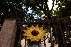 Sunflower design crocheted in circle handmade design yellow flower pattern made out of string yarn hung on metal fence wall barrier next to stone pillar in front of trees and building in Spanish town