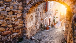 Stunning view of the street in old town of Ulcinj on sunset. Pots with flowers standing on paved pavement along stone wall. The sun light on stone arch. Travel Montenegro