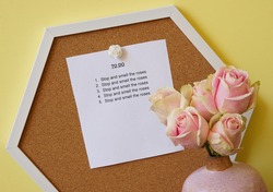 To Do list pinned to bulletin board against yellow painted wall says to stop and smell the roses.  Composition includes pale pink with light green edges roses in a vase.
