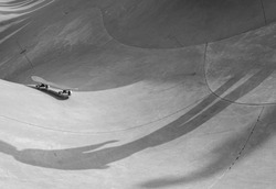 Black and white minimalist street photography shows shadow of person who lost his skateboard which is rolling away, against backdrop of curves of skate park.