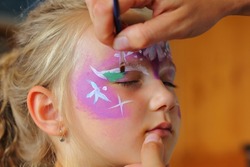 Children's face painting. The artist paints a princess crown on a small white blonde girl.