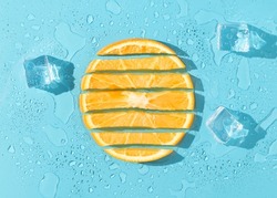 Orange fruit slices with melting ice cubes on blue background. Minimal citrus fruits concept. Creative summer refreshing food layout. Trendy vacation aesthetic. Flat lay with sunshine shadows.