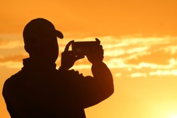 Silhouetted man back-lit by setting sun taking photo holding up a  mobile device