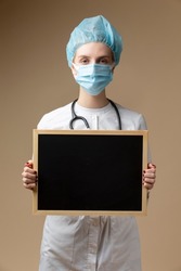 Young Professional Confident Female GP Doctor Posing in Doctor's Smock and Endoscope While Holding Black Board With Empty Space Over Beige. Vertical image