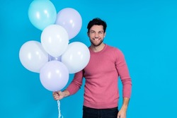 Celebration Concepts. Smiling Funny Caucasian Guy Handsome Brunet Man With Bunch of Colorful Air Balloons Posing in Pink Jumper Against Blue Background. Horizontal Image