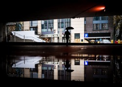 Lone worker man walking through underpass with umbrella up commuting to work in modern city centre flood water puddle reflected. Rainy day reflection dark silhouette bad severe weather big puddle