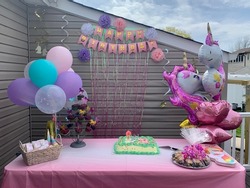 The close up view of a children's birthday party set up. The theme is unicorns with balloons, a banner, tissue paper flowers, cupcakes, cookies, plates, napkins, and an ice cream cake with candles.