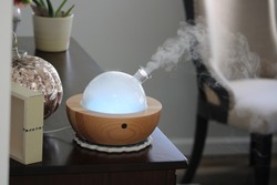 An oil diffuser that is on and producing a thick, steady stream of mist. It's diffusing essential oils while playing music and changing colors. The round remote is next to the machine.