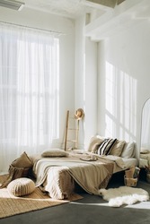 bedroom interior with white walls and large windows with white tulle. spacious bright bedroom filled with natural light. loft room with high ceilings and plenty of blankets and pillows on the bed.