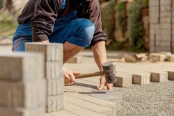 Young man laying gray concrete paving slabs in house courtyard on gravel foundation base. Master lays paving stones. Garden brick pathway paving by professional paver worker. Repairing sidewalk.
