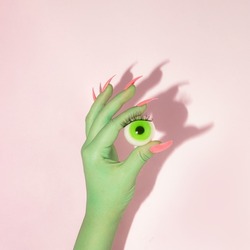 Creative layout with green painted hand with bright pink nails holding green eyeball against pastel pink background. Halloween celebration idea. Minimal flat lay concept.