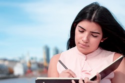 pretty latin girl, who is writing, taking notes in a black notebook with a black pen, in an urban industrial environment with the sky and buildings in the background. business and industrial concept.